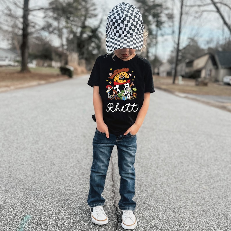 K&ouml;nnte beinhalten: Ein Kind tr&auml;gt ein schwarzes T-Shirt mit einem bunten Cartoon-Motiv und dem Namen "Rhett" in wei&szlig;er Schrift. Das Kind tr&auml;gt au&szlig;erdem blaue Jeans, wei&szlig;e Turnschuhe und eine schwarz-wei&szlig; karierte M&uuml;tze.