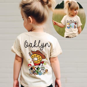 K&ouml;nnte beinhalten: Ein beiges T-Shirt mit dem Namen "Oaklyn" in schwarzer Schrift auf der R&uuml;ckseite. Das Shirt zeigt eine farbenfrohe Grafik von Toy Story-Charakteren, darunter Woody und Buzz Lightyear, mit einer gro&szlig;en "2".