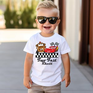 Puede incluir: Una camiseta blanca con un dise&ntilde;o de dibujos animados de dos personajes de Cars, Mate y Rayo McQueen, con banderas a cuadros. El texto "Two Fast Rhett" est&aacute; impreso debajo.