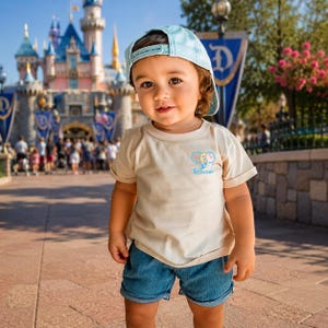 May include: A light beige short-sleeved t-shirt featuring a small graphic of a cartoon character and the text "Bro" on the chest, paired with a light blue baseball cap and blue denim shorts, worn by a toddler in a bright outdoor setting.