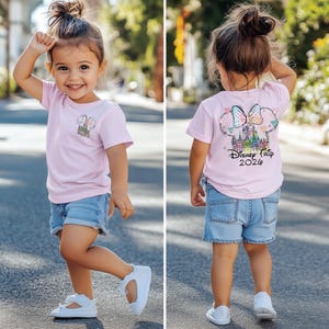 Pu&ograve; includere: Una bambina indossa una maglietta rosa chiaro e pantaloncini di jeans. La maglietta presenta una grafica colorata di un castello e la scritta "Disney Trip 2020". La bambina indossa anche scarpe bianche.