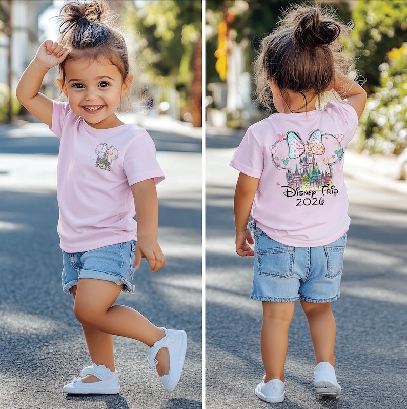 Pu&ograve; includere: Una bambina indossa una maglietta rosa chiaro e pantaloncini di jeans. La maglietta presenta una grafica colorata di un castello e la scritta "Disney Trip 2020". La bambina indossa anche scarpe bianche.