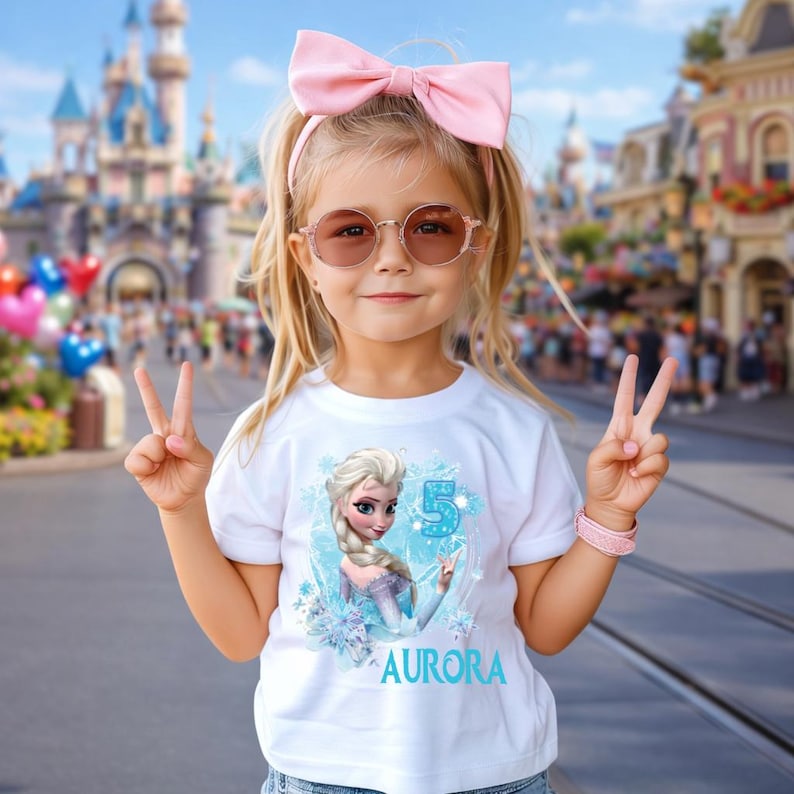 Pu&ograve; includere: Una bambina indossa una maglietta bianca con una grafica di Elsa di Frozen, il numero 5 e il nome "Aurora". Indossa un fiocco rosa, occhiali da sole e un braccialetto rosa. Lo sfondo mostra un parco a tema.