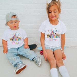 May include: Two children wearing white t-shirts with a colorful castle design and the words "Bro" and "Sister". The boy wears a gray cap, blue jeans, and gray sneakers. The girl wears denim shorts and white socks.