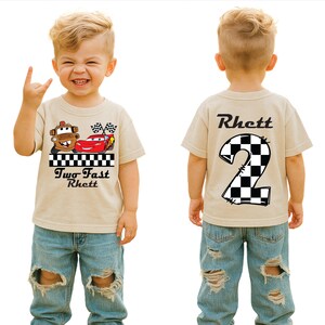 May include: A tan t-shirt featuring a cartoon car design with the text "Two Fast Rhett" on the front and a checkered number 2 on the back. The shirt is paired with blue denim jeans.