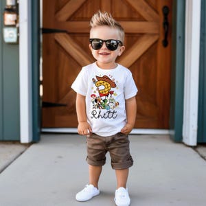 K&ouml;nnte beinhalten: Ein junges Kind tr&auml;gt ein wei&szlig;es T-Shirt mit einer bunten "Toy Story"-Grafik, einer gro&szlig;en "3" und dem Namen "Rhett". Das Kind tr&auml;gt au&szlig;erdem braune Shorts, wei&szlig;e Turnschuhe und eine schwarze Sonnenbrille. Der Hintergrund ist eine Holzt&uuml;r.