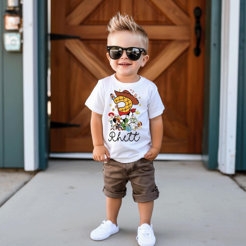 K&ouml;nnte beinhalten: Ein junges Kind tr&auml;gt ein wei&szlig;es T-Shirt mit einer bunten "Toy Story"-Grafik, einer gro&szlig;en "3" und dem Namen "Rhett". Das Kind tr&auml;gt au&szlig;erdem braune Shorts, wei&szlig;e Turnschuhe und eine schwarze Sonnenbrille. Der Hintergrund ist eine Holzt&uuml;r.