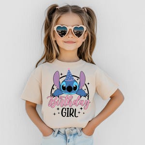May include: A beige t-shirt featuring a blue cartoon character with large ears and a party hat. The shirt has the text "Birthday GIRL" in pink and black. The girl is wearing heart-shaped sunglasses.