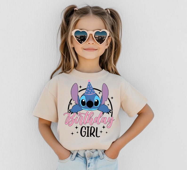 May include: A beige t-shirt featuring a blue cartoon character with large ears and a party hat. The shirt has the text "Birthday GIRL" in pink and black. The girl is wearing heart-shaped sunglasses.