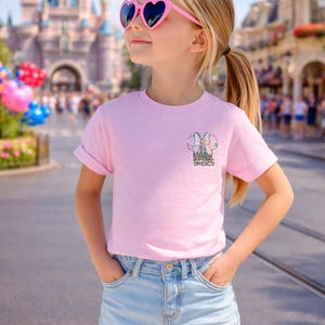 Pu&ograve; includere: Una persona giovane indossa una maglietta rosa chiaro con una grafica colorata e la scritta "Sadies". La persona indossa anche pantaloncini di jeans azzurri e occhiali da sole rosa a forma di cuore. Lo sfondo presenta un castello e una strada.
