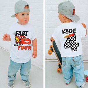 Pu&ograve; includere: Una t-shirt bianca a maniche corte con la grafica di un'auto da corsa rossa e la scritta FAST AND FOUR sul davanti, e il nome KADE sopra un grande numero 4 a scacchi sul retro, abbinata a un berretto da baseball grigio e jeans azzurri.