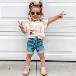 K&ouml;nnte beinhalten: Ein junges Kind tr&auml;gt ein cremefarbenes T-Shirt mit den Worten "Birthday Girl" in Pastellfarben und einer Grafik von zwei Zeichentrickfiguren. Das Kind tr&auml;gt au&szlig;erdem blaue Jeansshorts, eine Sonnenbrille und beige Schuhe.