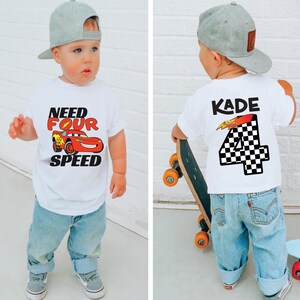 Op de afbeelding: Een wit T-shirt met korte mouwen voorzien van een rode raceauto-afbeelding met de tekst "NEED FOUR SPEED" op de voorzijde en de naam "KADE" boven een groot geblokt cijfer "4" op de achterzijde, gecombineerd met een lichtblauwe jeans en een grijze pet.