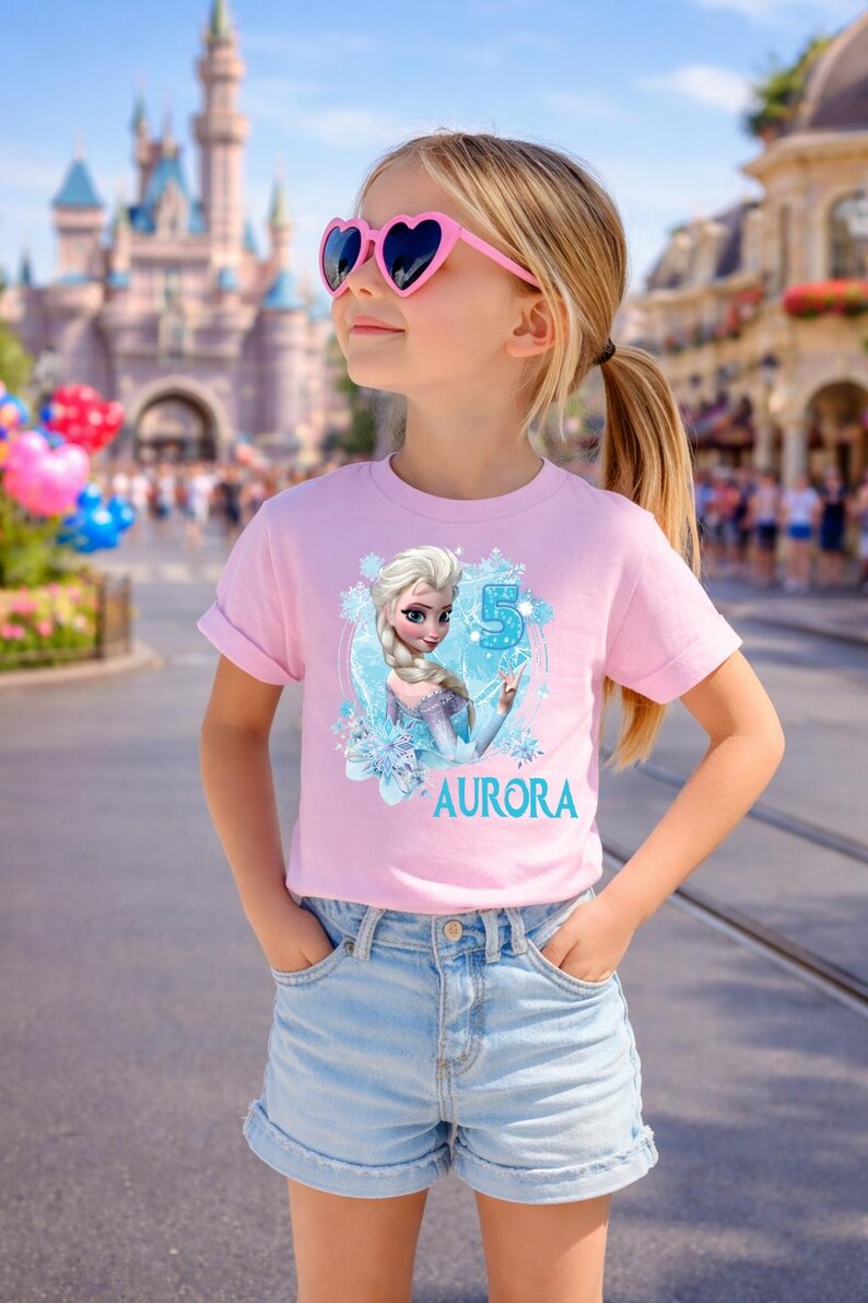 Pu&ograve; includere: Una bambina indossa una maglietta rosa con un personaggio dei cartoni animati e il nome "AURORA". La maglietta presenta anche il numero "5". Indossa pantaloncini di jeans e occhiali da sole rosa a forma di cuore.