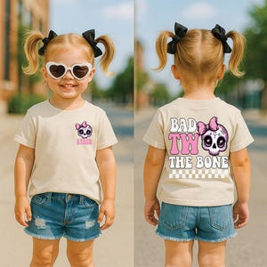 May include: A beige t-shirt featuring a small skull graphic with a pink bow on the front left chest with the name Ryder below it. The back displays a large skull with a pink bow and the text Bad To The Bone with a checkered pattern below.