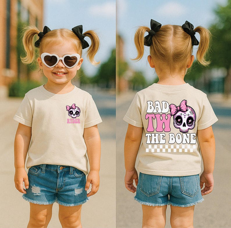 May include: A beige t-shirt featuring a small skull graphic with a pink bow on the front left chest with the name Ryder below it. The back displays a large skull with a pink bow and the text Bad To The Bone with a checkered pattern below.