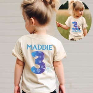 K&ouml;nnte beinhalten: Beiges T-Shirt mit dem Namen "MADDIE" und der Zahl "3" in Blau und Lila, verziert mit Schneeflocken. Auf der Vorderseite des Shirts steht "3 BIRTHDAY GIRL" mit einem Bild von Zeichentrickfiguren.