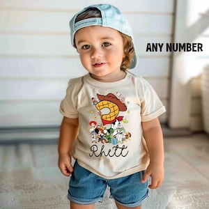 K&ouml;nnte beinhalten: Ein Kleinkind tr&auml;gt ein hellbeige T-Shirt mit einer bunten Grafik mit der Zahl 2 und Figuren aus Toy Story. Auf dem Shirt steht der Name "Rhett" unter der Grafik. Das Kind tr&auml;gt au&szlig;erdem blaue Jeansshorts und eine hellblaue Baseballkappe.