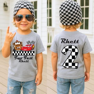 May include: A gray t-shirt with a race car design and the text "Two Fast Rhett" on the front. The back of the shirt features a large number "2" in a checkered pattern and the name "Rhett". The child is wearing a checkered beanie and sunglasses.