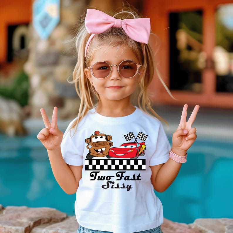 Puede incluir: Una ni&ntilde;a peque&ntilde;a con una camiseta blanca con un gr&aacute;fico de dos coches de dibujos animados y el texto "Two Fast Sissy". Tambi&eacute;n lleva gafas de sol rosas y una diadema rosa. Est&aacute; haciendo el signo de la paz.