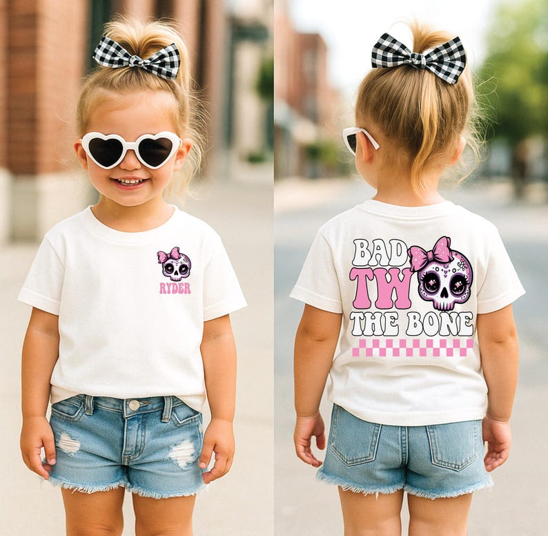 May include: A white short-sleeve t-shirt featuring a small pink skull graphic with a bow on the front chest with the name Ryder. The back displays a large pink skull with a bow, the text Bad To The Bone, and a pink and white checkered pattern.