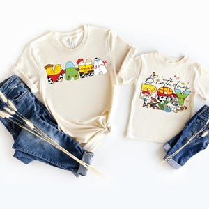 May include: Two cream-colored t-shirts with the words "MAMA" and "Birthday Boy" printed on them in colorful, cartoon-style letters. The shirts are paired with blue jeans and decorative dried flowers.