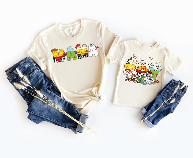 May include: Two cream-colored t-shirts with the words "MAMA" and "Birthday Boy" printed on them in colorful, cartoon-style letters. The shirts are paired with blue jeans and decorative dried flowers.