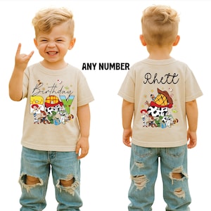 May include: A tan short-sleeved t-shirt with a colorful graphic featuring characters and the text "Birthday Boy." The back of the shirt has the name "Rhett" and a similar graphic. The shirt is worn by a child.