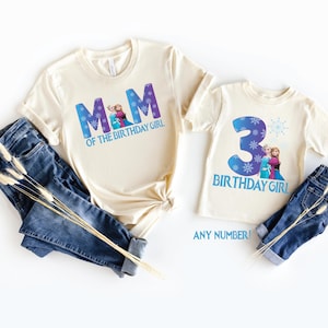 K&ouml;nnte beinhalten: Zwei cremefarbene T-Shirts mit blauem Text und Grafiken. Auf einem Shirt steht "MOM OF THE BIRTHDAY GIRL" und auf dem anderen "3 BIRTHDAY GIRL". Beide Shirts sind mit blauen Jeans kombiniert. Der Text "ANY NUMBER!" ist ebenfalls sichtbar.
