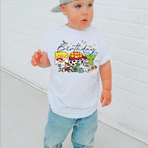 May include: A white t-shirt with a colorful "Birthday Boy" graphic featuring Toy Story characters. The child is wearing light blue jeans, gray sneakers, and a gray baseball cap. The background is a white brick wall.