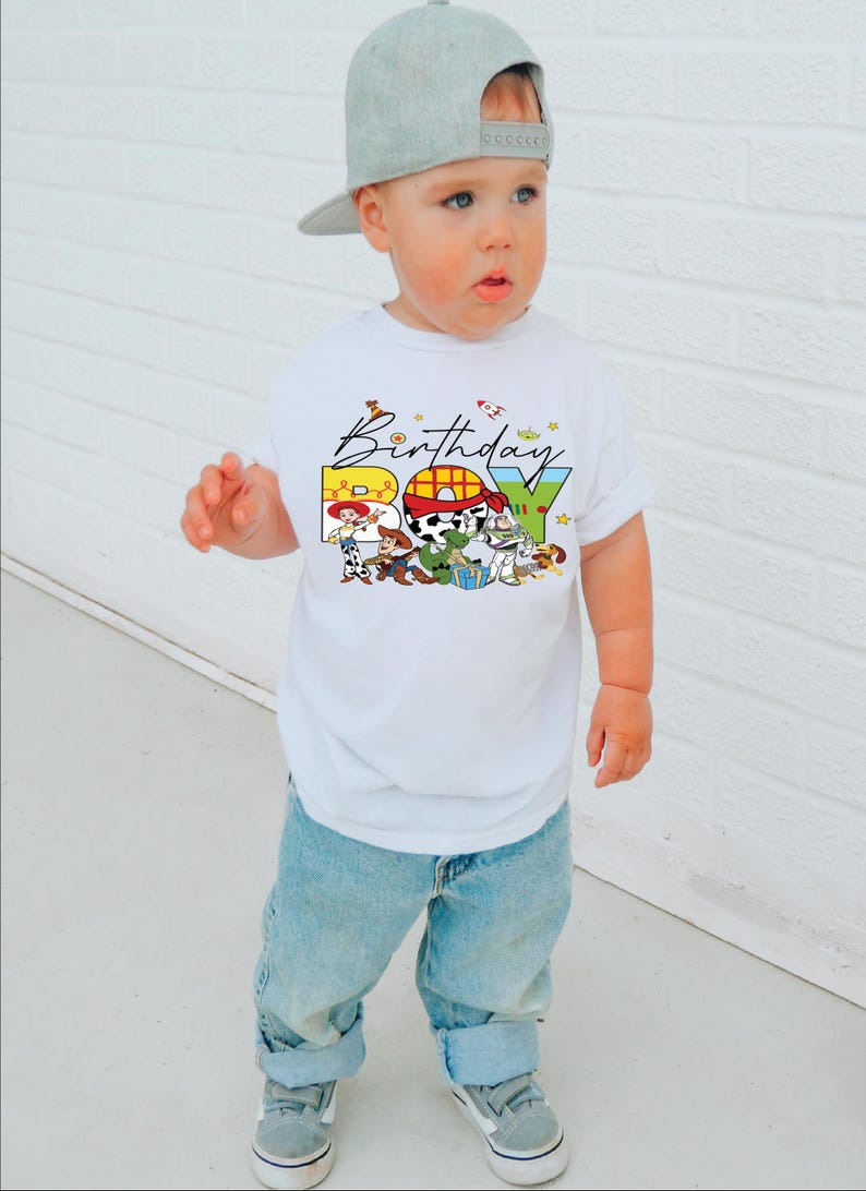 May include: A white t-shirt with a colorful "Birthday Boy" graphic featuring Toy Story characters. The child is wearing light blue jeans, gray sneakers, and a gray baseball cap. The background is a white brick wall.