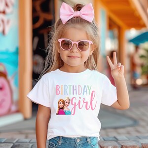 K&ouml;nnte beinhalten: Ein junges M&auml;dchen tr&auml;gt ein wei&szlig;es T-Shirt mit den Worten "BIRTHDAY girl" in Pastellfarben und einer Grafik von zwei Zeichentrickfiguren. Sie tr&auml;gt auch eine rosa Sonnenbrille und eine gro&szlig;e rosa Schleife in ihrem Haar.