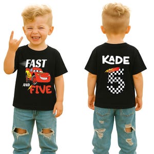 K&ouml;nnte beinhalten: Ein schwarzes Kurzarm-T-Shirt mit einer roten Rennwagen-Grafik und dem Text "FAST AND FIVE" auf der Vorderseite sowie dem Namen "KADE" &uuml;ber einer gro&szlig;en karierten Zahl "5" auf der R&uuml;ckseite, kombiniert mit einer hellblauen Jeans.