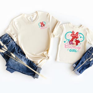 May include: Two cream-colored t-shirts and two pairs of blue jeans. The larger shirt features a small graphic of Ariel with a red bow and the word Mama. The smaller shirt features a large graphic of Ariel with a red bow and the text Birthday Girl.