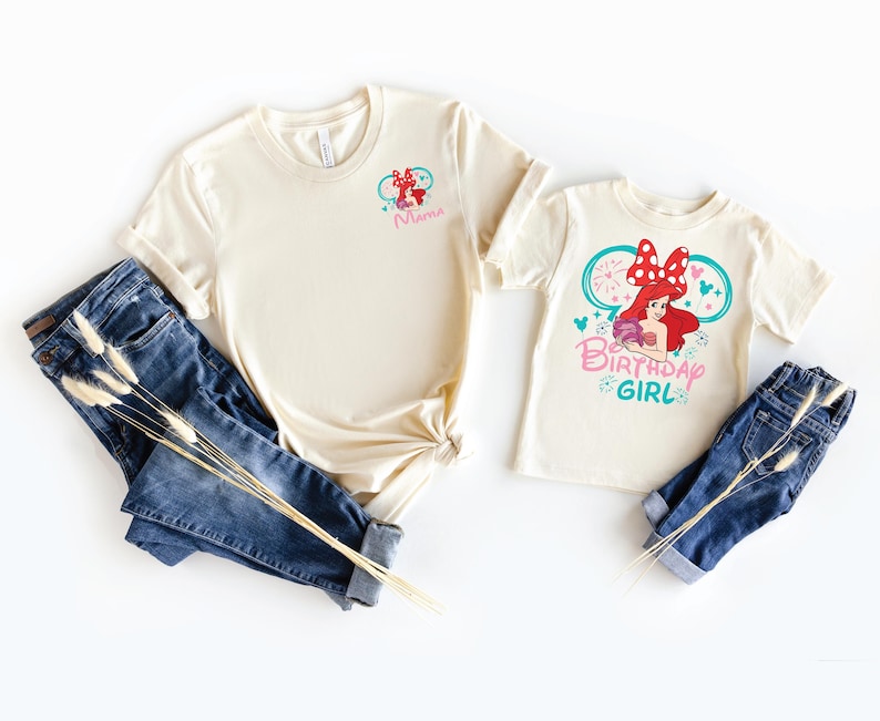 May include: Two cream-colored t-shirts and two pairs of blue jeans. The larger shirt features a small graphic of Ariel with a red bow and the word Mama. The smaller shirt features a large graphic of Ariel with a red bow and the text Birthday Girl.