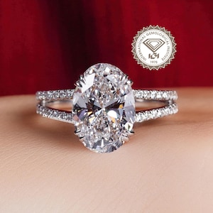 May include: An oval-cut diamond engagement ring with a split band, set with smaller diamonds. The ring is displayed against a red background, with a logo in the upper right corner. The ring is a symbol of love and commitment.