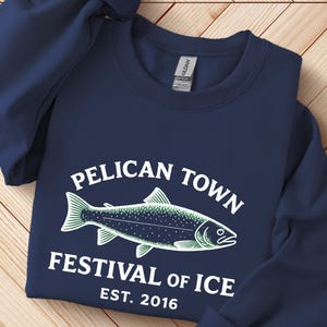 以下が含まれることがあります： 「Pelican Town Festival of Ice Est. 2016」の文字と魚のグラフィックが白と緑で描かれたネイビーブルーのスウェットシャツ。明るい茶色の木製の表面に折りたたまれています。