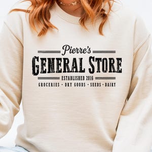 以下が含まれることがあります： ヴィンテージの黒いフォントで「Pierre's GENERAL STORE」と書かれたクリーム色のスウェットシャツ。デザインには設立年2016年と、店の名前の下に「GROCERIES • DRY GOODS • SEEDS • DAIRY」が記載されています。