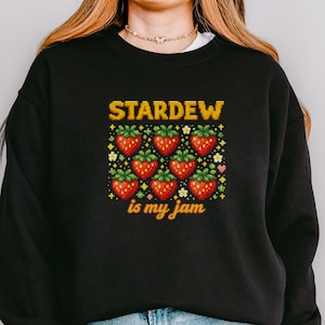 以下が含まれることがあります： 「STARDEW is my jam」の文字が黄色と白でプリントされた黒いスウェットシャツ。デザインは、緑の葉が付いた8つの赤いイチゴのグリッドが特徴です。スウェットシャツは青いジーンズと合わせて着用されています。