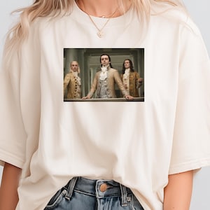 May include: Cream-colored t-shirt featuring a rectangular image of three people in historical attire. The image shows three men in ornate jackets and vests, standing behind a railing. The t-shirt is paired with denim shorts.