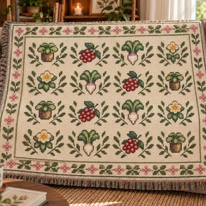 May include: A woven throw blanket with a cream-colored base featuring a repeating grid pattern of potted plants, red berries, white radishes, and yellow flowers. The blanket is framed by a border of pink flowers and green leaves with fringed edges.
