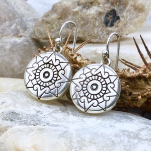 May include: A pair of silver dangle earrings with a circular design featuring a stylised sun with a central eye. The earrings have a simple hook fastening.
