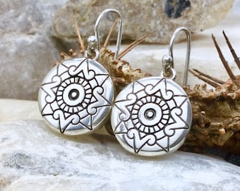 May include: A pair of silver dangle earrings with a circular design featuring a stylized sun with a central eye. The earrings have a simple hook closure.