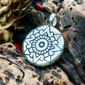 May include: A silver pendant featuring a detailed design of a stylised sun with a central eye. The pendant is round and has a textured surface.