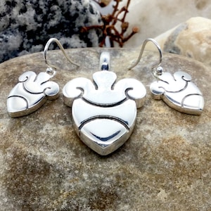 May include: A silver pendant and matching earrings set featuring a heart-shaped design with intricate details. The pendant and earrings have a smooth, polished finish.