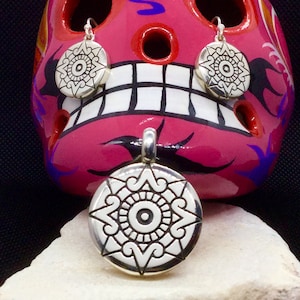 May include: A set of silver earrings and a pendant featuring a stylised sun design with a central eye. The jewellery is displayed on a pink and white painted skull.