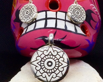 May include: A set of silver earrings and a pendant featuring a stylized sun design with a central eye. The jewelry is displayed on a pink and white painted skull.