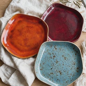 Peut inclure: Trois assiettes en céramique de forme carrée avec des coins arrondis et des bords irréguliers. Une assiette est orange vif, une autre est rouge bordeaux profond, et la troisième est bleu clair avec des taches brun foncé sur la glaçure.