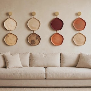 Op de afbeelding: Vier verticale wanddecoraties, elk bestaande uit twee onregelmatige, bordachtige vormen die aan een ronde gouden bevestiging hangen. De borden vertonen diverse aardetinten, waaronder crème, beige, donkerrood en terracotta met texturen.