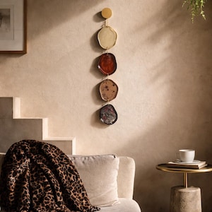 Puede incluir: Un colgante de pared con cuatro platos de forma irregular en crema, rojo, marrón y gris oscuro. Los platos están suspendidos de un disco dorado. La pared es de color beige neutro. Una manta con estampado de leopardo está sobre un sofá.